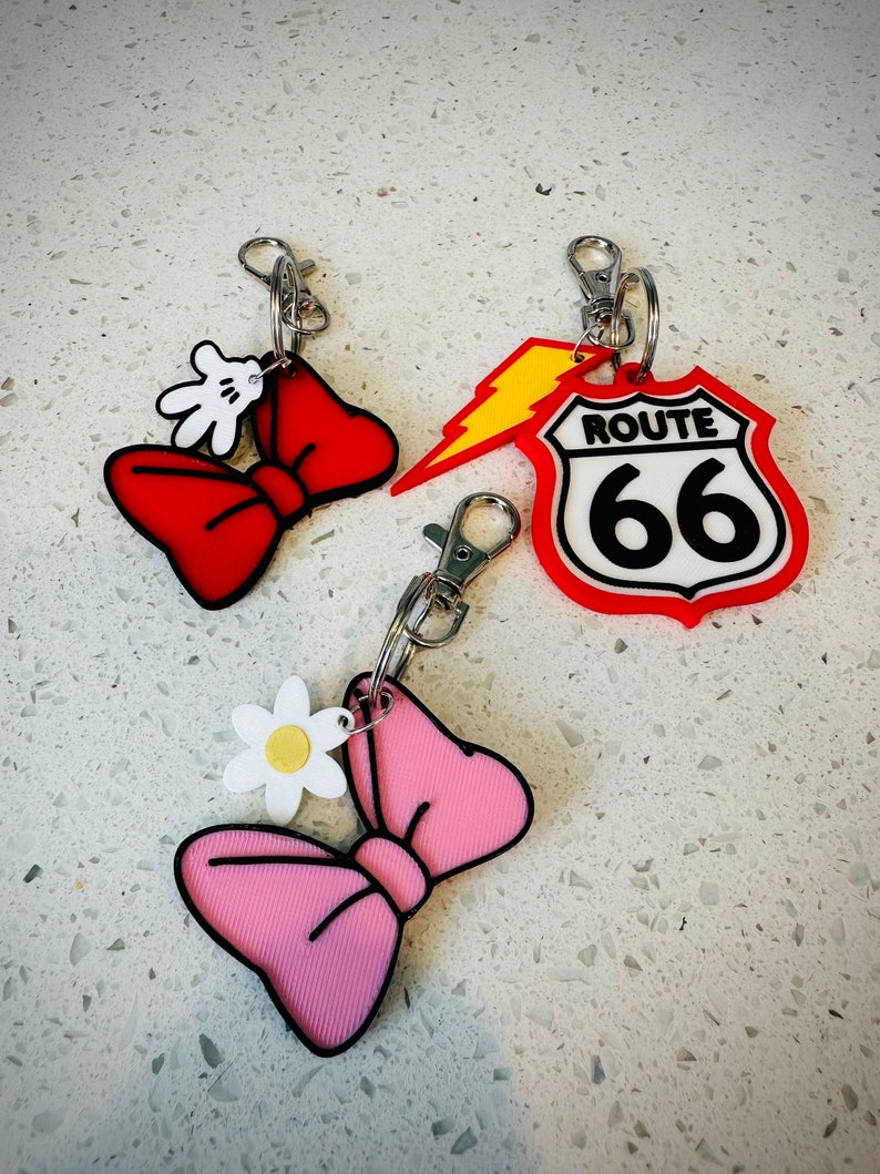 May include: Three keychains with different designs. One is a red bow with a white Mickey Mouse hand, another is a pink bow with a white daisy, and the last is a red Route 66 sign with a yellow lightning bolt.