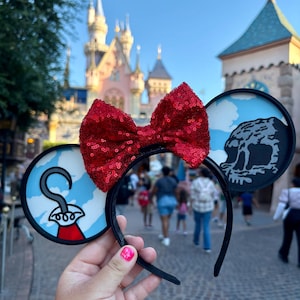 May include: Black headband with red sequined bow and two circular ear designs. One ear features a skull and clouds, the other a cartoon hook and red coat design. The background shows a castle and people walking.
