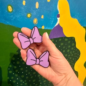 May include: Two lavender-colored bow-shaped accessories with black outlines. The bows are held in front of a colorful background depicting a blue sky with floating lanterns and a yellow-haired figure. The bows are likely decorative items or accessories.
