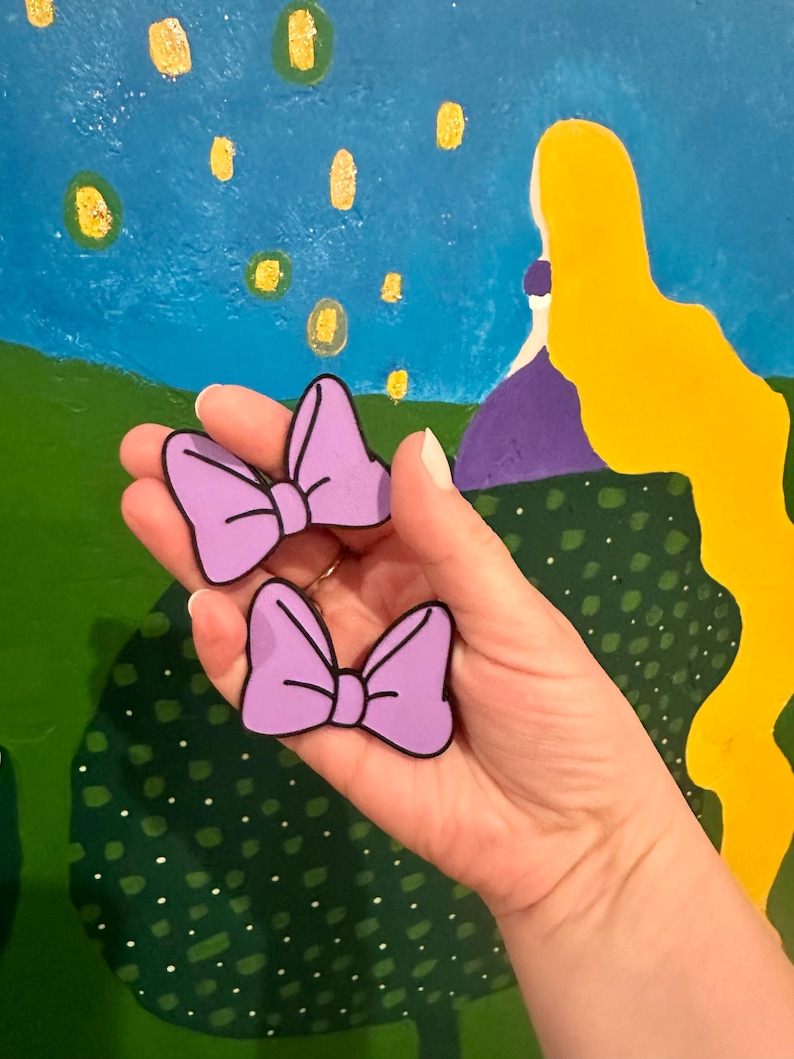 May include: Two lavender-colored bow-shaped accessories with black outlines. The bows are held in front of a colorful background depicting a blue sky with floating lanterns and a yellow-haired figure. The bows are likely decorative items or accessories.
