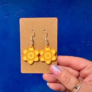 May include: A pair of yellow and orange atom-shaped earrings with silver hooks. The earrings are displayed on a brown card against a blue background. The earrings have a science theme.