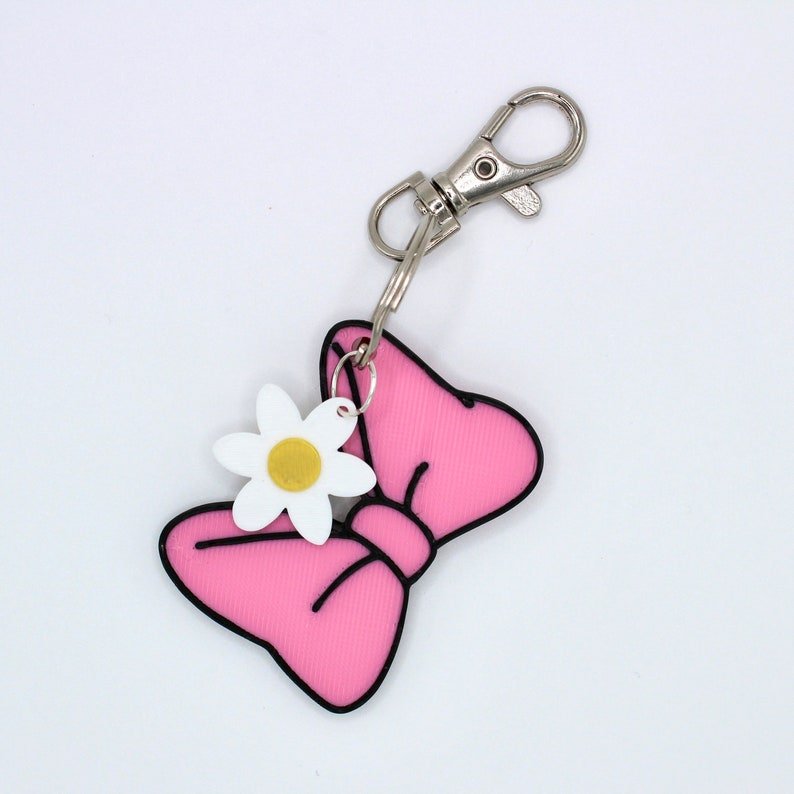 May include: Pink bow keychain with a white flower charm. The bow has a black outline and the flower has a yellow center.