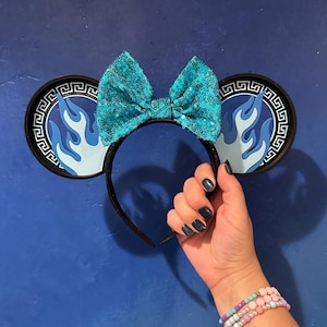 May include: Black headband with round ears featuring a blue flame design and a Greek key border. A large, sequined turquoise bow sits atop the headband. The ears are held by a hand with dark blue nail polish, and a beaded bracelet is visible.