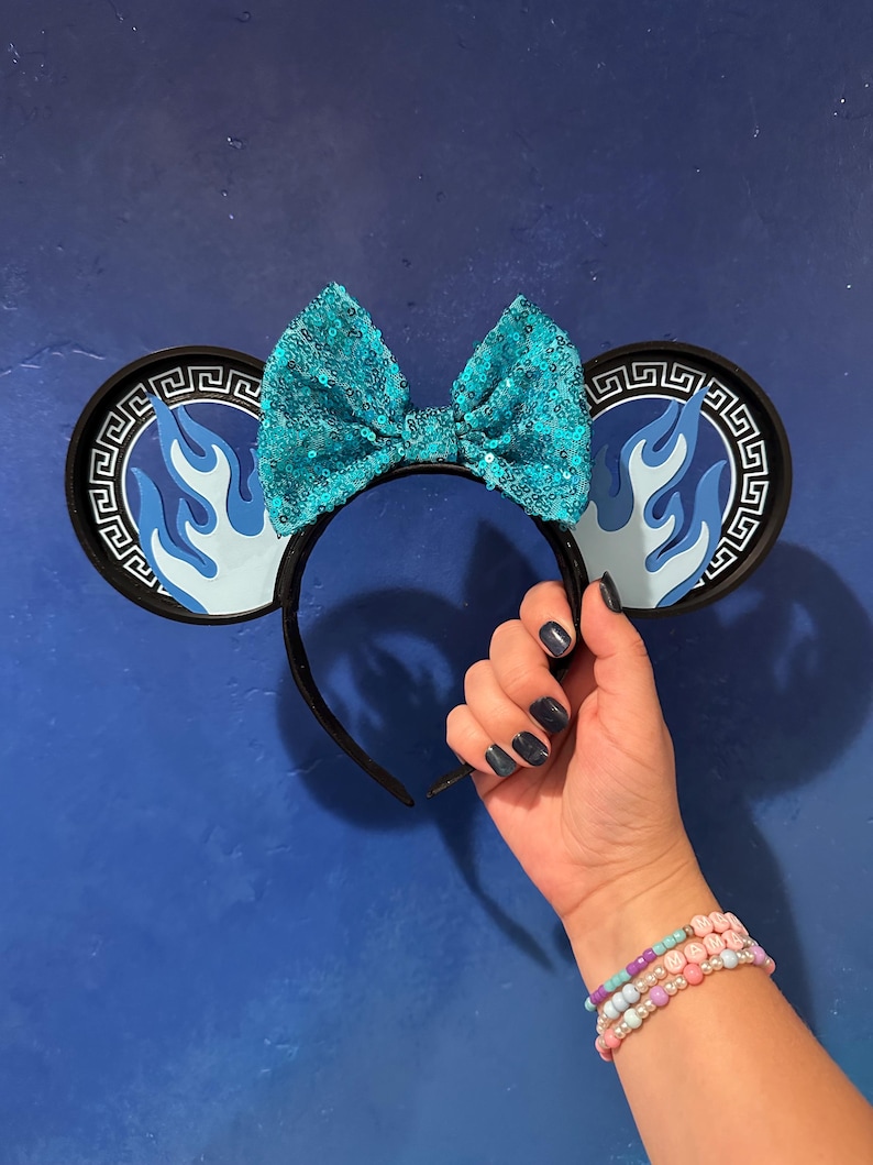 May include: Black headband with round ears featuring a blue flame design and a Greek key border. A large, sequined turquoise bow sits atop the headband. The ears are held by a hand with dark blue nail polish, and a beaded bracelet is visible.