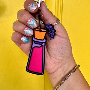 May include: A keychain featuring a flat, stylized potion bottle charm with a bright pink center, a purple border, and an orange stopper. Attached to the silver metal lobster clasp is a small, dark purple feather-shaped charm.