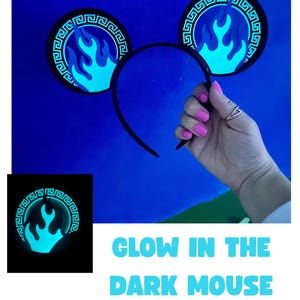 May include: Black mouse ear headband with glowing blue flame design. The ears have a Greek key border. The text "GLOW IN THE DARK MOUSE EARS OPTION!" is displayed in blue. The ears are held up by a hand with pink nail polish.