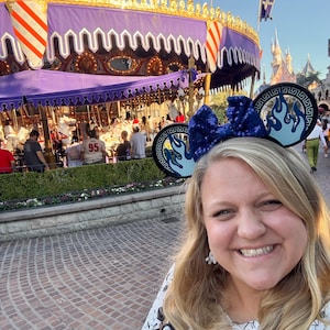 May include: A woman wearing a blue sequined bow headband with flame designs, posing in front of a carousel. The carousel has a purple canopy and is decorated with gold trim. The background includes a castle and other park attractions.