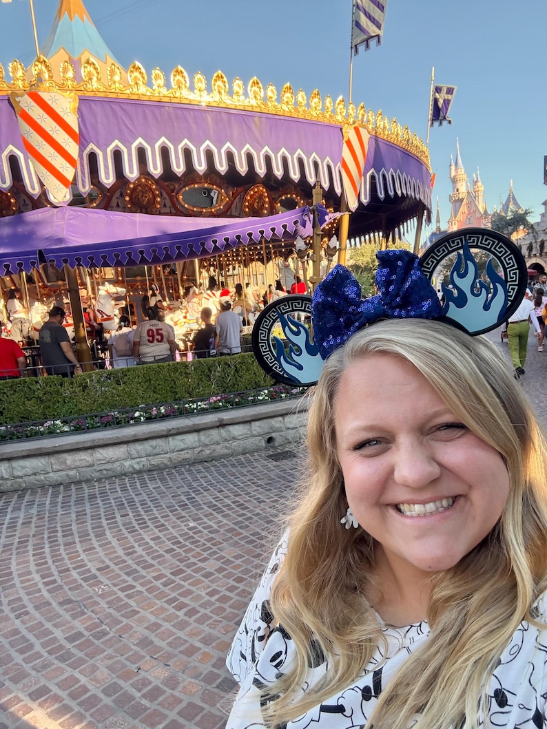 May include: A woman wearing a blue sequined bow headband with flame designs, posing in front of a carousel. The carousel has a purple canopy and is decorated with gold trim. The background includes a castle and other park attractions.