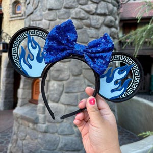 May include: Black headband with round ears featuring a blue flame design and a Greek key border. A large, sequined blue bow sits atop the headband. The ears are held up by a black headband.
