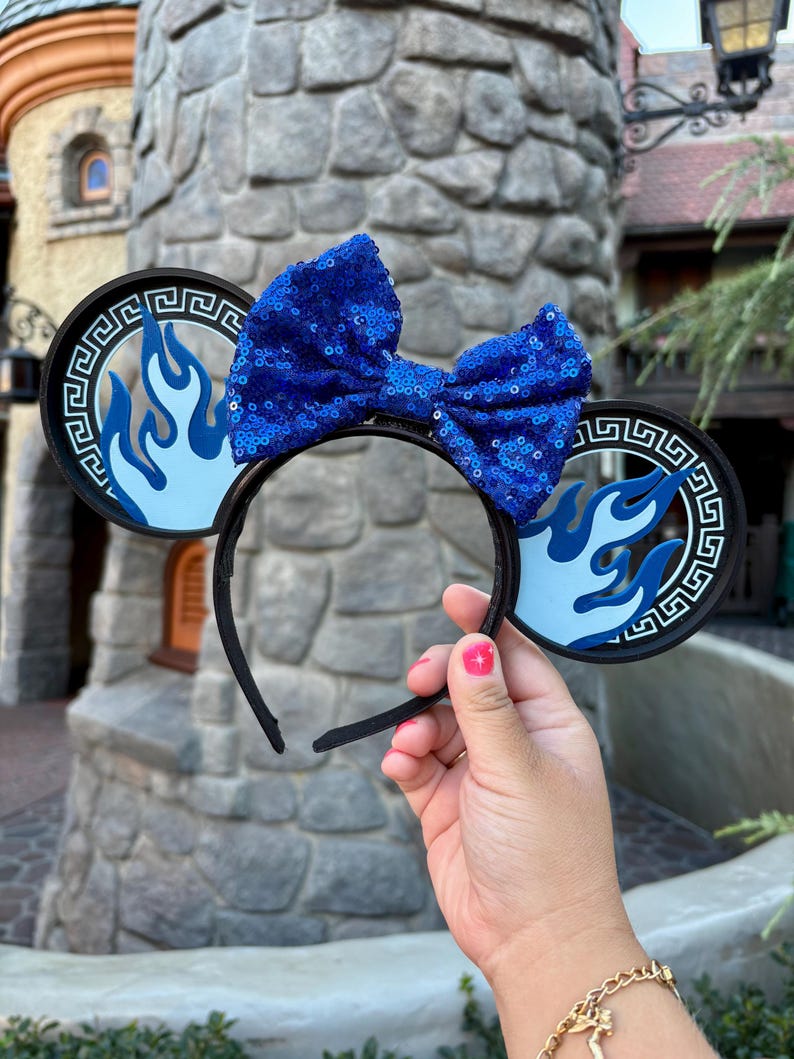 May include: Black headband with round ears featuring a blue flame design and a blue sequin bow. The ears have a black border with a Greek key pattern. The headband is held up by a person.