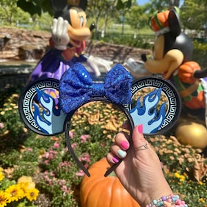 May include: A black headband with blue sequined bow and round ears. The ears have a blue flame design and a Greek key border. The headband is held in front of a pumpkin and flower garden with Mickey Mouse statues in the background.