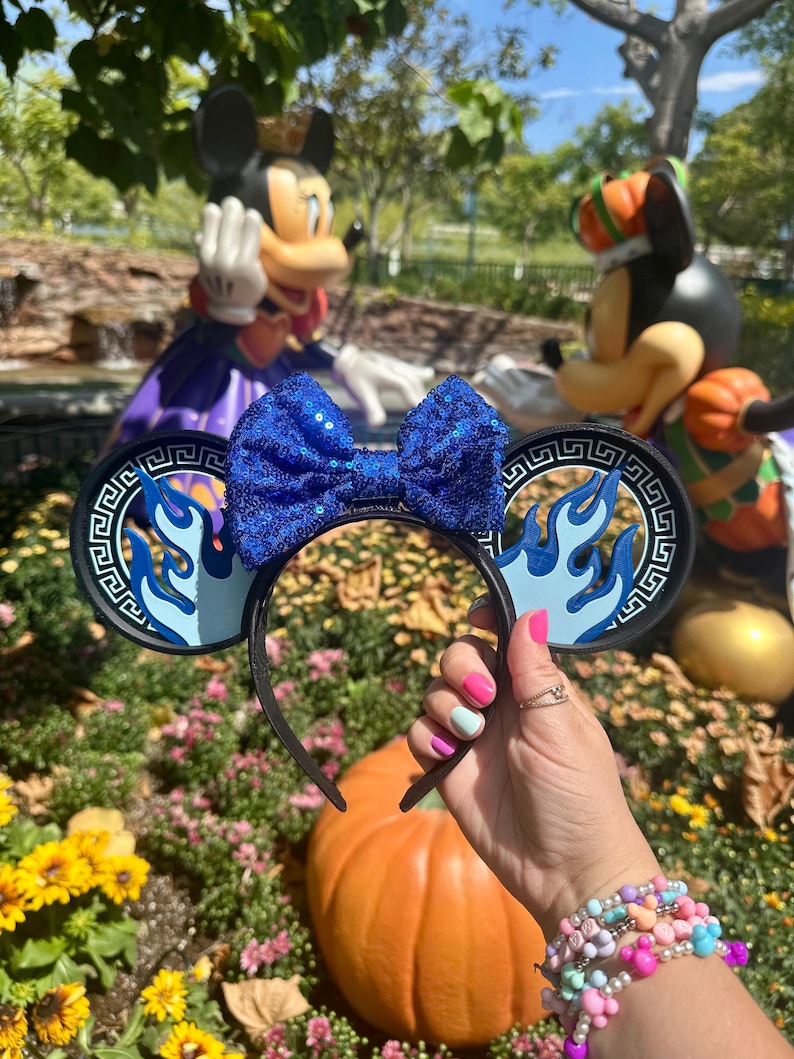 May include: A black headband with blue sequined bow and round ears. The ears have a blue flame design and a Greek key border. The headband is held in front of a pumpkin and flower garden with Mickey Mouse statues in the background.