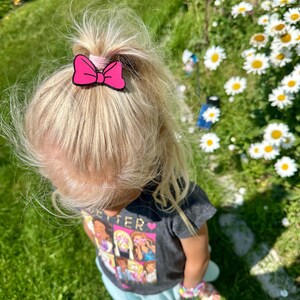 May include: A child's blonde hair is pulled up in a ponytail, secured with a pink bow hair clip. The child is wearing a gray t-shirt with a colorful graphic and a light blue skirt, standing on green grass.