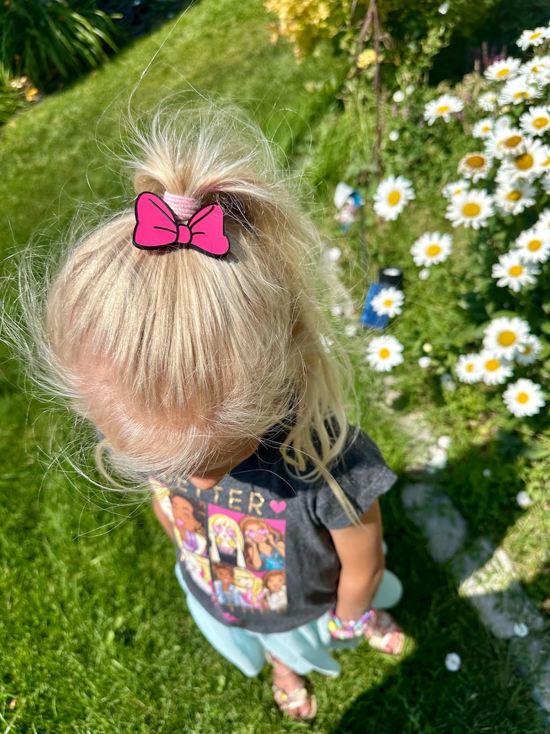 May include: A child's blonde hair is pulled up in a ponytail, secured with a pink bow hair clip. The child is wearing a gray t-shirt with a colorful graphic and a light blue skirt, standing on green grass.