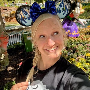 May include: A woman wearing a black t-shirt and a pair of Mickey Mouse ears with a blue sequined bow and flame design. A Minnie Mouse statue is in the background, along with a garden and trees.