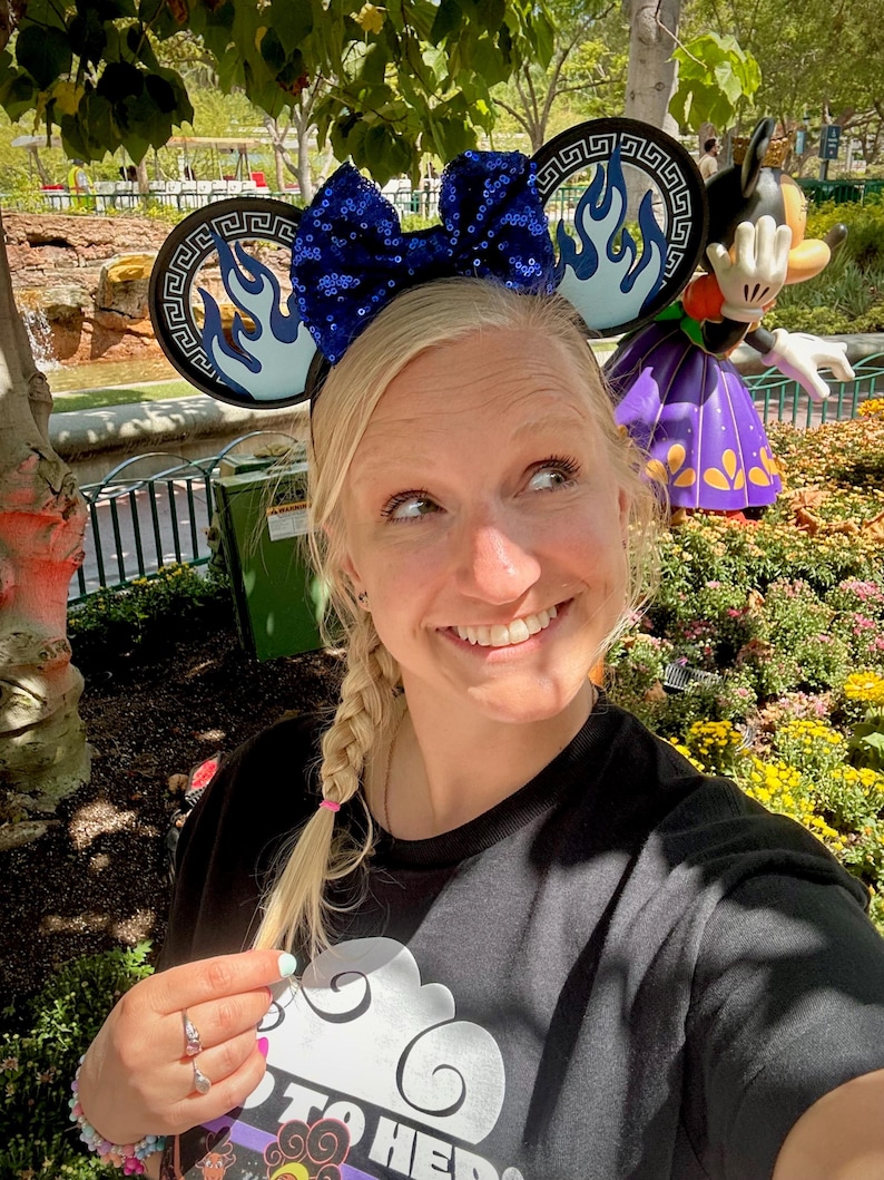 May include: A woman wearing a black t-shirt and a pair of Mickey Mouse ears with a blue sequined bow and flame design. A Minnie Mouse statue is in the background, along with a garden and trees.