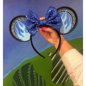 May include: A black headband with attached mouse ears. The ears feature a blue flame design and a decorative border. A large, sequined blue bow is centered on the headband. The text "GLOW IN THE DARK EARS" is at the bottom.