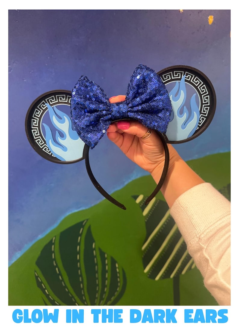 May include: A black headband with attached mouse ears. The ears feature a blue flame design and a decorative border. A large, sequined blue bow is centered on the headband. The text "GLOW IN THE DARK EARS" is at the bottom.