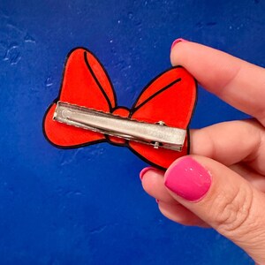 May include: A red bow-shaped hair clip with a black outline and a silver metal clip on the back. The bow is held by a hand with pink nail polish against a blue background.