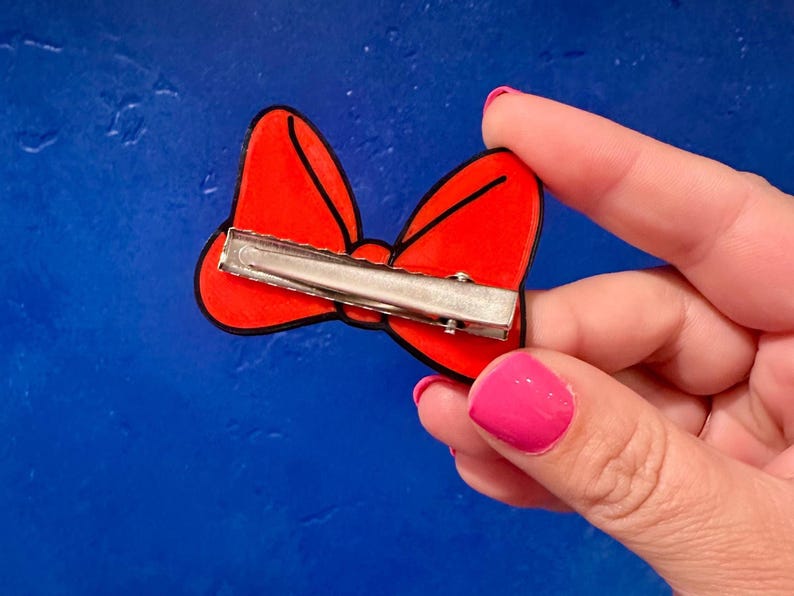 May include: A red bow-shaped hair clip with a black outline and a silver metal clip on the back. The bow is held by a hand with pink nail polish against a blue background.