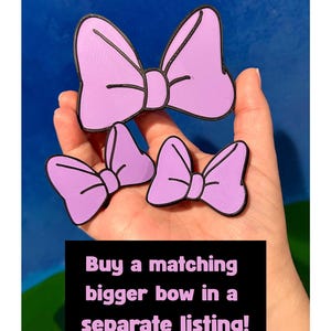 May include: Three lavender-colored bow-shaped accessories with black outlines. The bows are in different sizes. Text on a black background reads: "Buy a matching bigger bow in a separate listing!"
