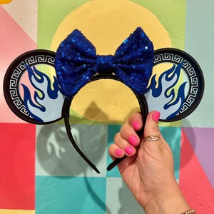May include: Black headband with attached ears featuring a blue flame design and a sequined blue bow. The ears have a Greek key border. The headband is held up by a hand with pink nail polish.