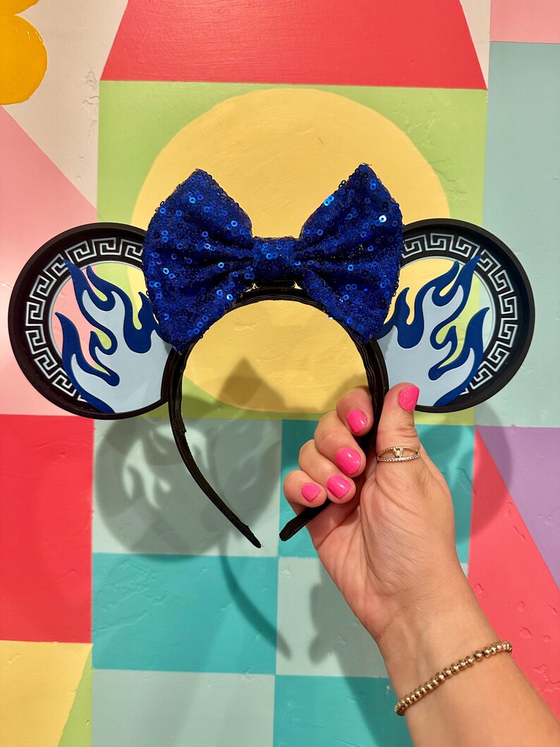 May include: Black headband with attached ears featuring a blue flame design and a sequined blue bow. The ears have a Greek key border. The headband is held up by a hand with pink nail polish.