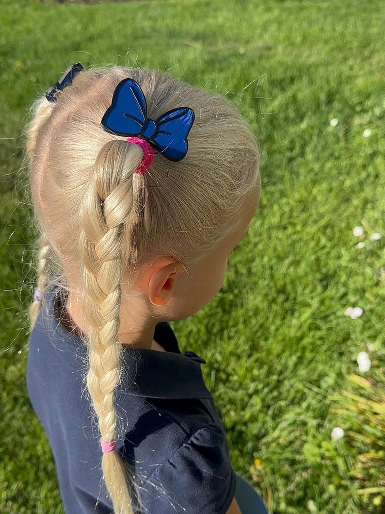 May include: A young child with blonde hair styled in braids and pigtails. A blue bow hair clip is attached to the hair with a pink hair tie. The child is wearing a navy blue shirt, and the background is green grass.