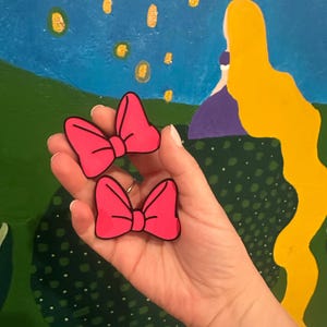 May include: Two bright pink bow-shaped accessories with black outlines are held in a hand. The bows are a vibrant fuchsia color. The background features a painted scene with a blue sky, green landscape, and yellow accents.