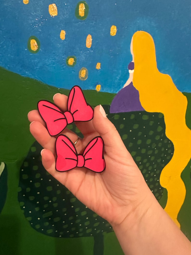 May include: Two bright pink bow-shaped accessories with black outlines are held in a hand. The bows are a vibrant fuchsia color. The background features a painted scene with a blue sky, green landscape, and yellow accents.