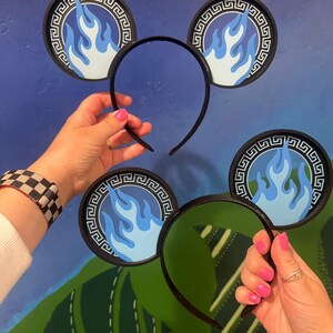 May include: Black headband with circular mouse ears featuring a blue flame design. The ears have a black border with a Greek key pattern. The text "NORMAL MOUSE EARS" and "GLOW IN THE DARK MOUSE EARS" are visible.