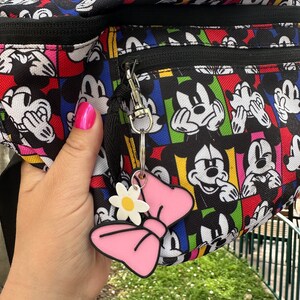 May include: A black fanny pack with a colorful Mickey Mouse print. The pack has a zipper closure and a pink bow keychain with a white flower.