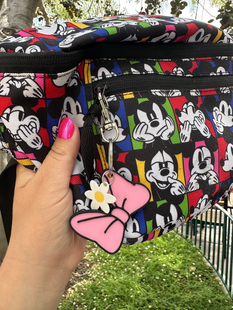 May include: A black fanny pack with a colorful Mickey Mouse print. The pack has a zipper closure and a pink bow keychain with a white flower.