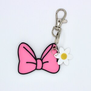 May include: Pink bow keychain with a white flower charm. The bow has a black outline and the flower has a yellow center.