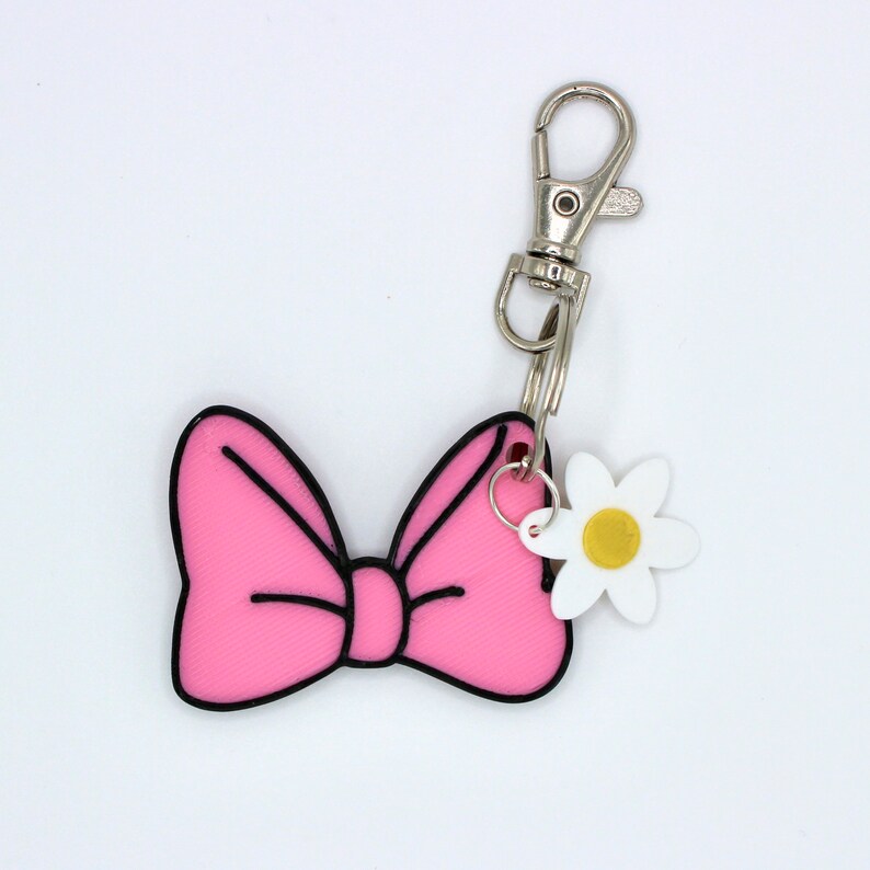 May include: Pink bow keychain with a white flower charm. The bow has a black outline and the flower has a yellow center.