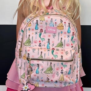 May include: A pink backpack with a Disney Princesses print. The backpack features a variety of princesses, including Cinderella, Belle, Ariel, and Tiana. The backpack has a zipper closure and a small pocket on the front. The backpack also has a pink bow charm attached to the zipper.