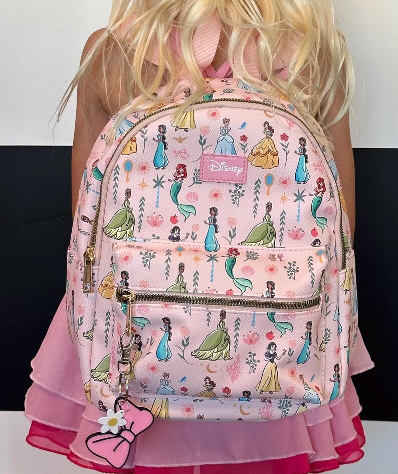 May include: A pink backpack with a Disney Princesses print. The backpack features a variety of princesses, including Cinderella, Belle, Ariel, and Tiana. The backpack has a zipper closure and a small pocket on the front. The backpack also has a pink bow charm attached to the zipper.