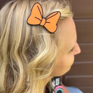 May include: An orange bow hair clip with a black outline is attached to blonde hair. The bow is a solid orange color and is in the shape of a classic bow. The hair clip is a simple accessory.