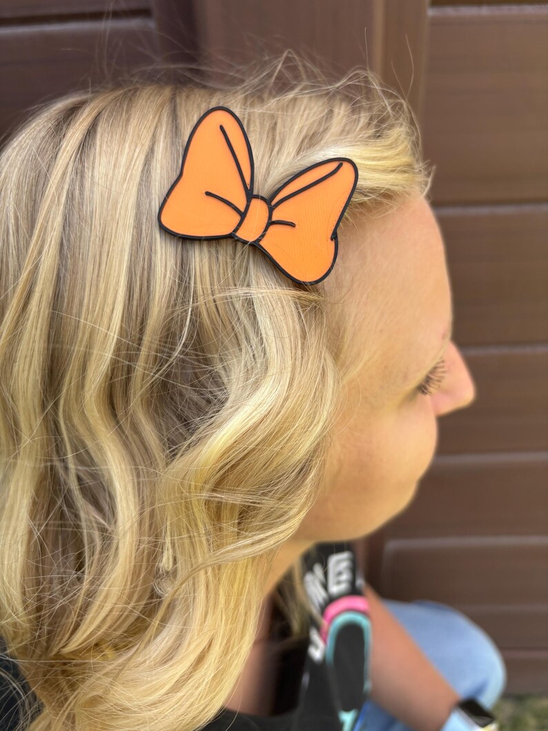 May include: An orange bow hair clip with a black outline is attached to blonde hair. The bow is a solid orange color and is in the shape of a classic bow. The hair clip is a simple accessory.
