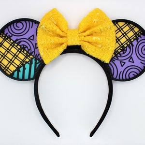 May include: Black headband with two round ears featuring a patchwork design in yellow, purple, and teal. A large, yellow sequined bow sits between the ears. The ears have a black border.