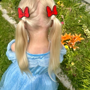 A young child with blonde hair styled in pigtails, each with a red bow-shaped hair clip.