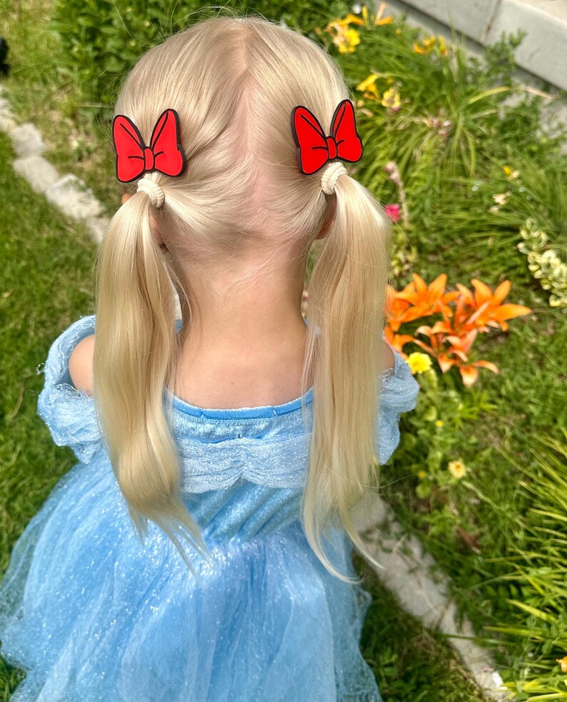 A young child with blonde hair styled in pigtails, each with a red bow-shaped hair clip.
