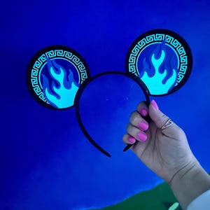 May include: Black headband with two circular ear decorations. Each ear features a blue flame design within a black border with a Greek key pattern. The ears are held by a hand with pink nail polish against a blue background.