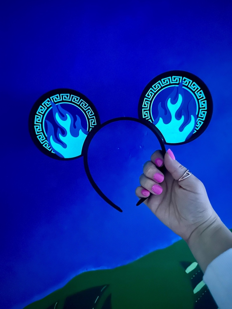 May include: Black headband with two circular ear decorations. Each ear features a blue flame design within a black border with a Greek key pattern. The ears are held by a hand with pink nail polish against a blue background.