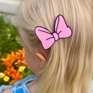 A pink bow-shaped hair clip with a black outline is clipped into blonde hair.