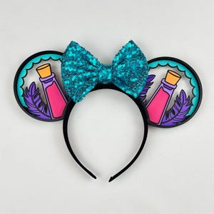 May include: A black headband featuring two circular ears with a teal scalloped border. Each ear displays a pink potion bottle with a tan cork and a purple feather. A large, bright teal sequined bow is centered between the two ears.