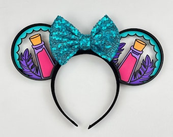 May include: A black headband featuring two circular ears with a teal sequined bow in the center. Each ear displays a graphic of a pink potion bottle with a tan cork and purple feathers against a teal scalloped background and black frame.