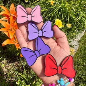 May include: Four colorful bow-shaped accessories in pink, purple, and red, each with a black outline. The bows are held in a hand, with a bracelet visible on the wrist. The background features green foliage and orange flowers.