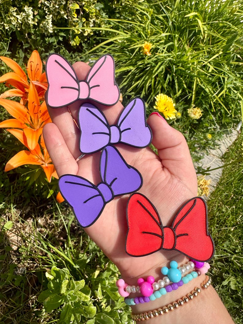 May include: Four colorful bow-shaped accessories in pink, purple, and red, each with a black outline. The bows are held in a hand, with a bracelet visible on the wrist. The background features green foliage and orange flowers.