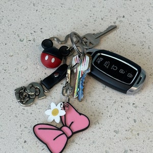 May include: A set of keys with a pink bow keychain, a white daisy charm, a silver Mickey Mouse charm, and a black car key with a silver key ring.